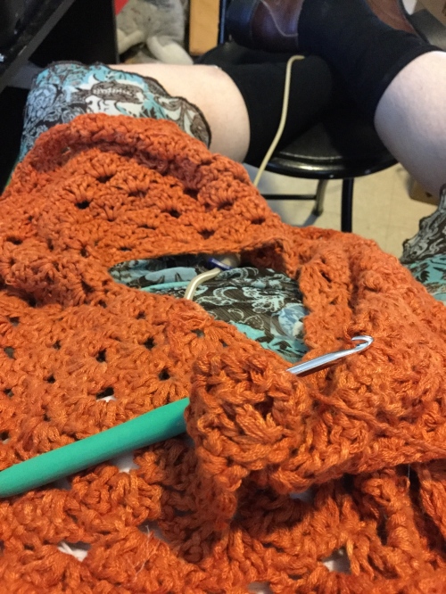 An orange crochet project sitting on Mel's lap, bamboo yarn with a lot of shell stitches that is going to become a cardigan, with a metal crochet hook with a green handle.